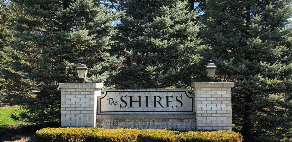 Brick pillars with lanterns and sign reading 'The Shires', in front of pine trees