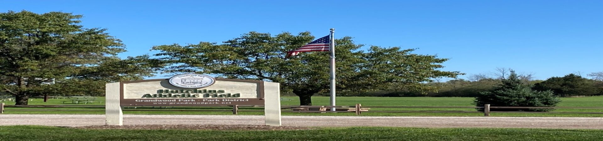 picture of a field with a white and brown park district sign and a flagpole and trees and bushes