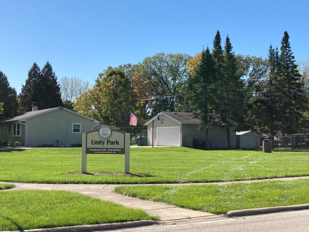 picture of a field with a white and brown park district sign and trees with houses in the background