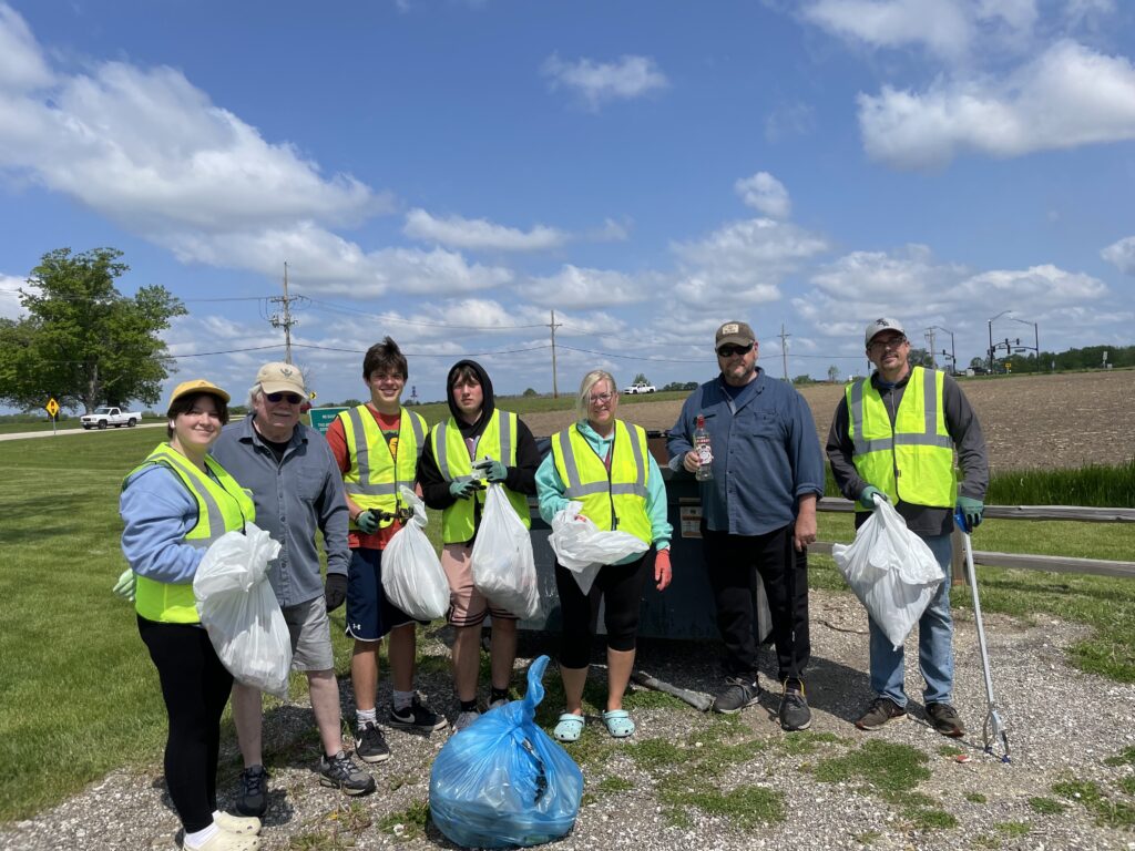 7 volunteers for Adopt a Highway cleanup standing in front of a fence