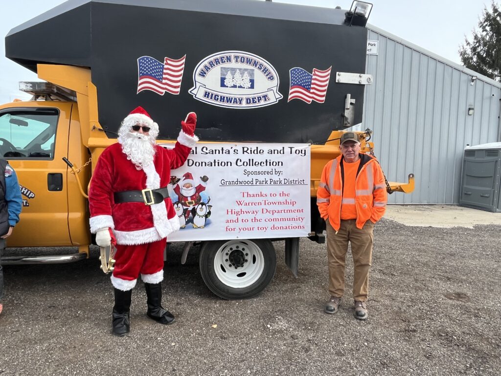 Santa in a red & white suit with another man in an orange jacket in front of a yellow truck